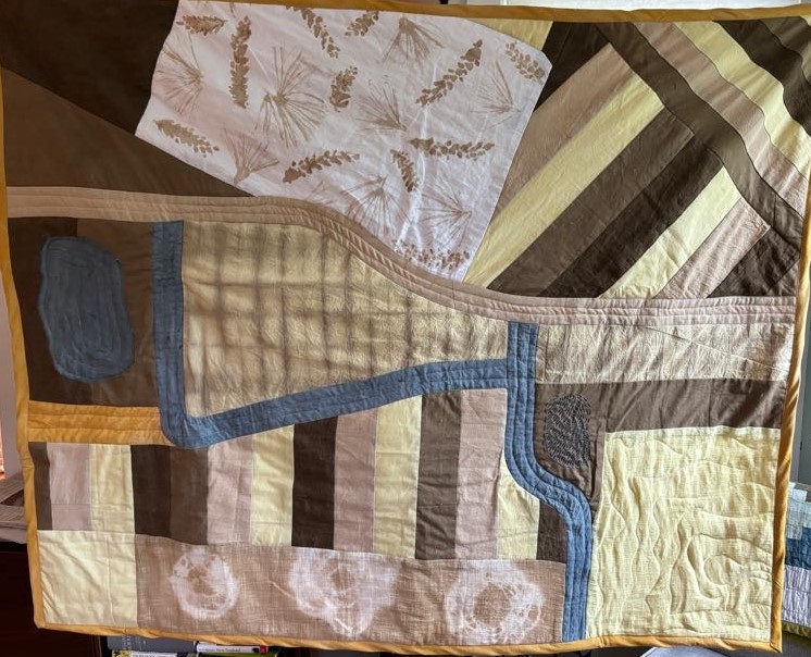 A 40 by 50 inch quilt representing an aerial view of the North Woods, including trails, water features, and plant communities. The fabric is dyed yellow, various browns, blue, and gold.